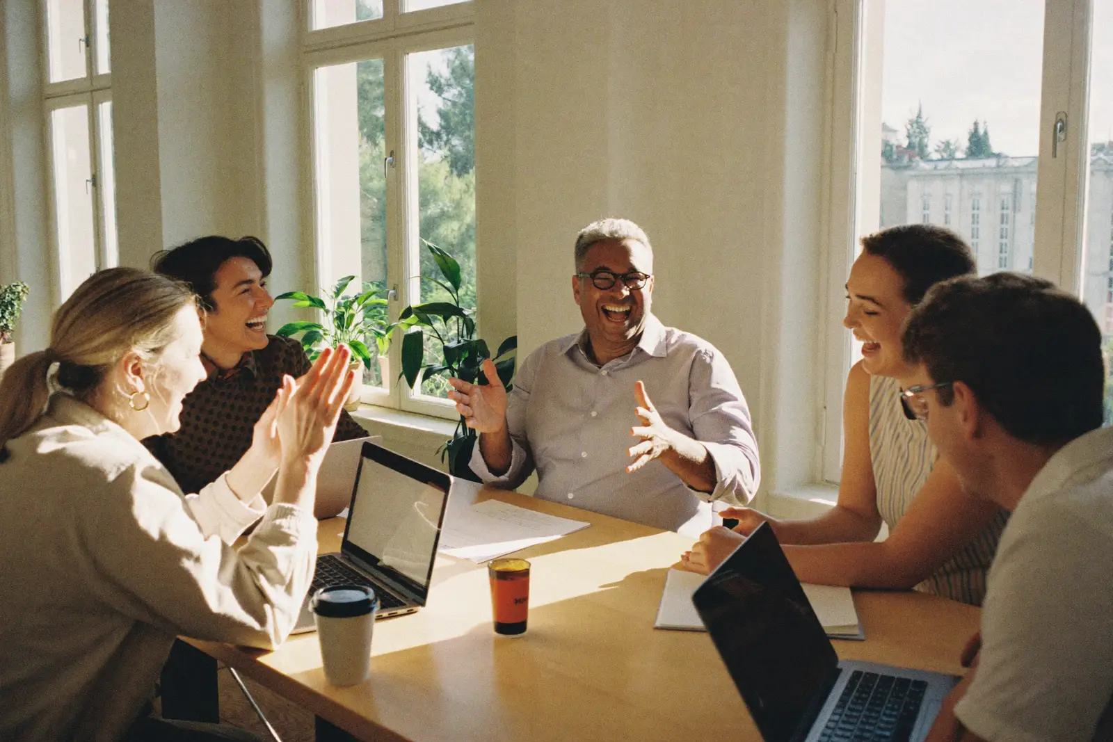 Team lacht gemeinsam am Besprechungstisch im Sonnenlicht